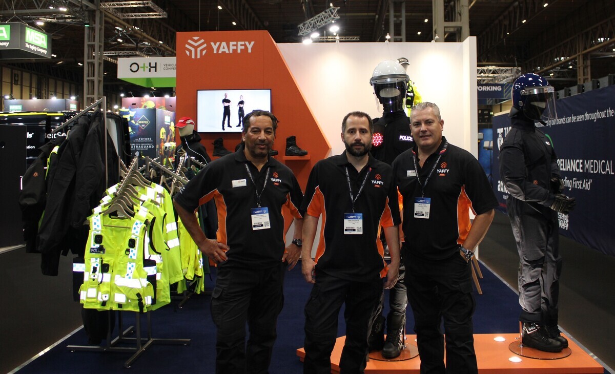 Three people stood in front of an exhibition stand with uniform garments on display on mannequins and clothes rails. 