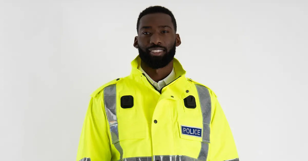 A close up of a Police Officer wearing a hi-visibility Police jacket stood in front of a plain white background.